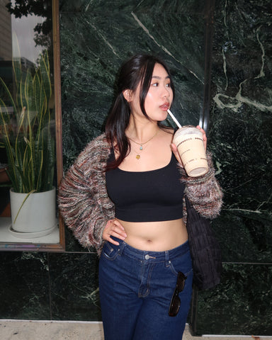 Person holding a coffee drink posing in front of a green marble wall with a black top and a fuzzy cardigan