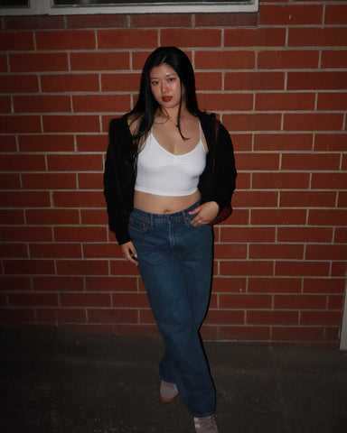 Woman standing against a brick wall wearing a black jacket, white top, and blue jeans.
